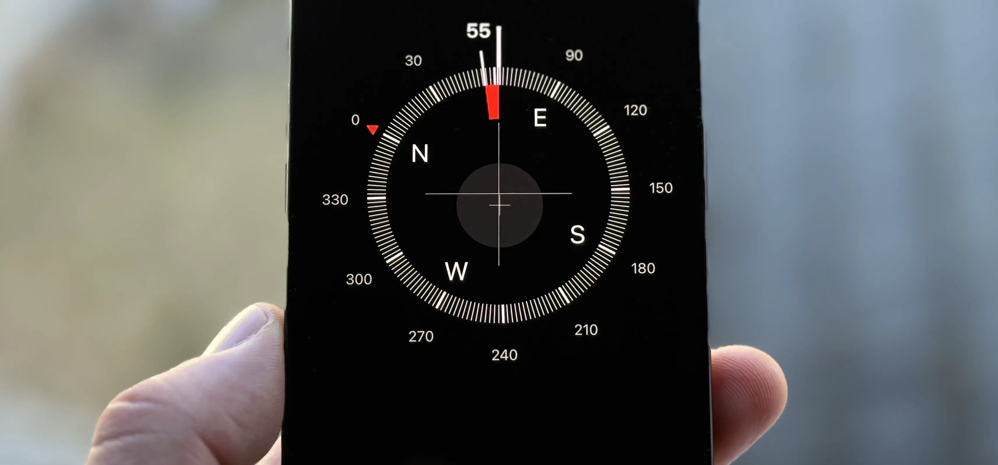A hand holds a smartphone displaying a digital compass app, showing a heading of 55 degrees northeast. The background is blurred with neutral tones.