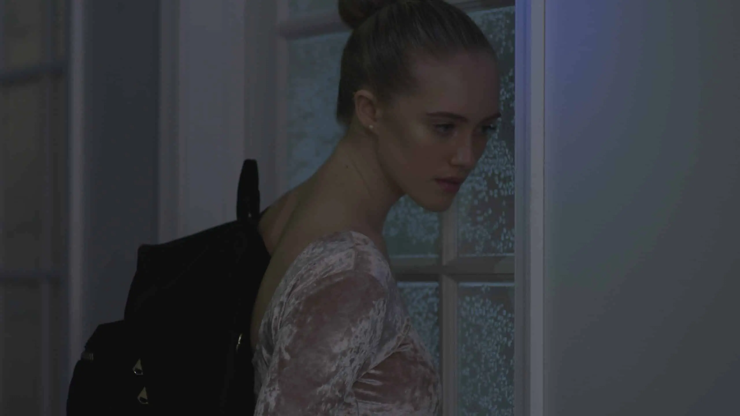 Pretty Vulgar 4 Dimmed image of a female with short blond hair in a bun wearing a black backpack looking offsides by a paned window.