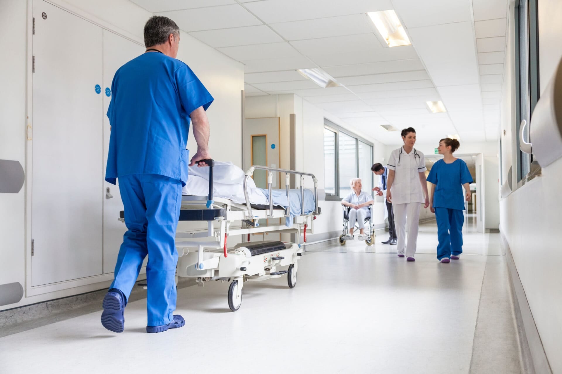 Male nurse pushing stretcher gurney bed in hospital corridor with doctors & senior female patient