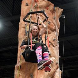 A woman wearing funky pink and purple socks is scaling an adaptive climbing wall using the Wellman climbing chair.
