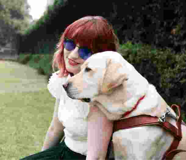 A person and a yellow Guide Dog, in harness, sitting on grass outside. The person is smiling at the camera and the Guide Dog is a profile view.
