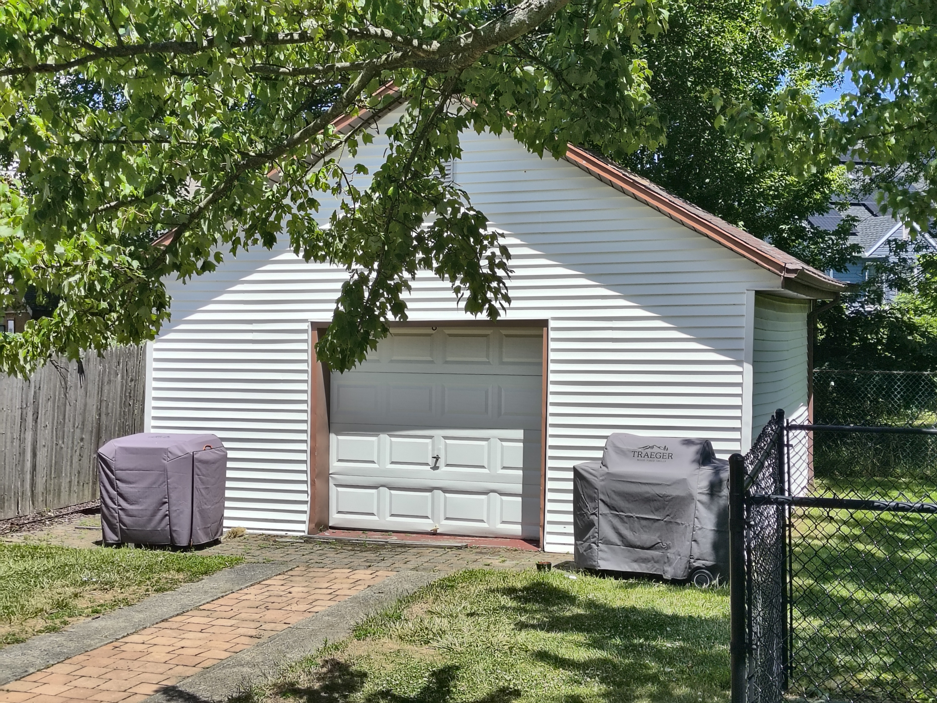 White wooden garage surrounded by trees and green grass with two outdoor chairs under weather-proof covers in the sun