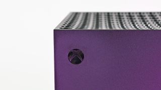 Macro shot of the circular Xbox logo power button on the corner of the console, under a purple metallic sheen.