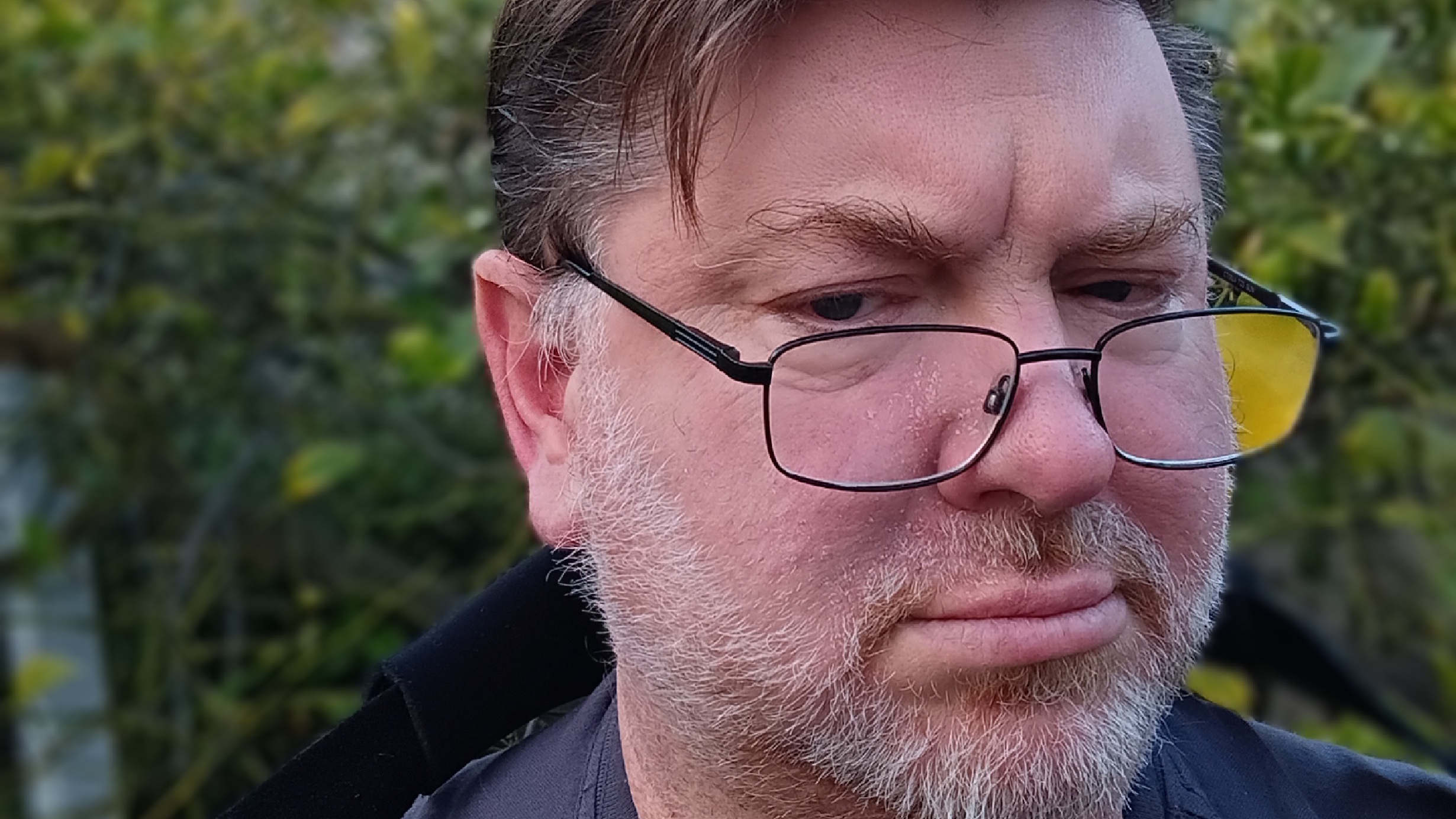Philip Michaels wearing glasses slid to the end of his nose taking a selfie outside in front of a green bush