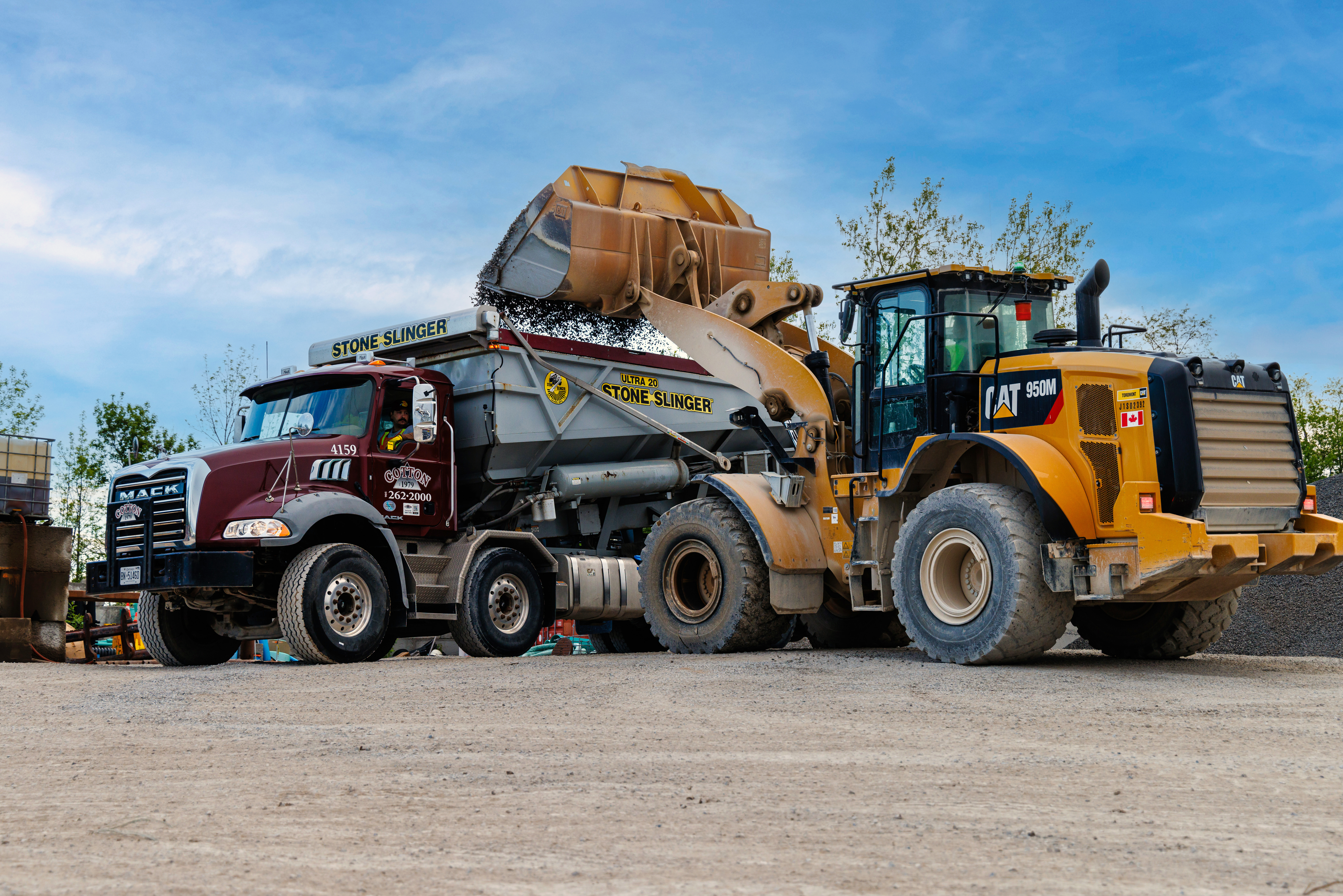 This image shows a large back hoe dumping gravel into a Cotton Inc stone slinger truck. 