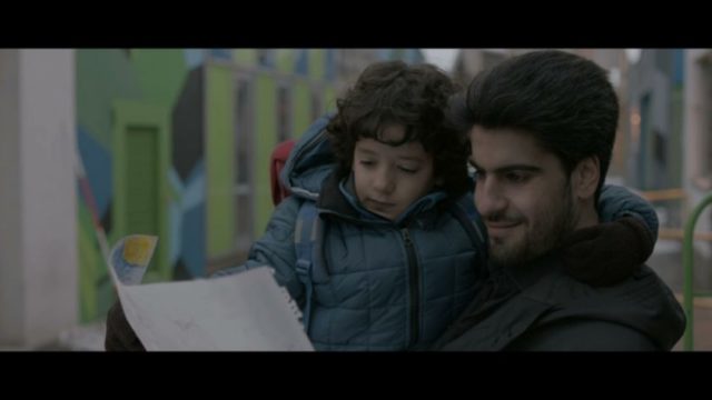 Fotograma de Crípticamente en el que aparece un hombre con un niño pequeño en brazos. Sujeta una hoja de papel y sonríe mientras la observa.