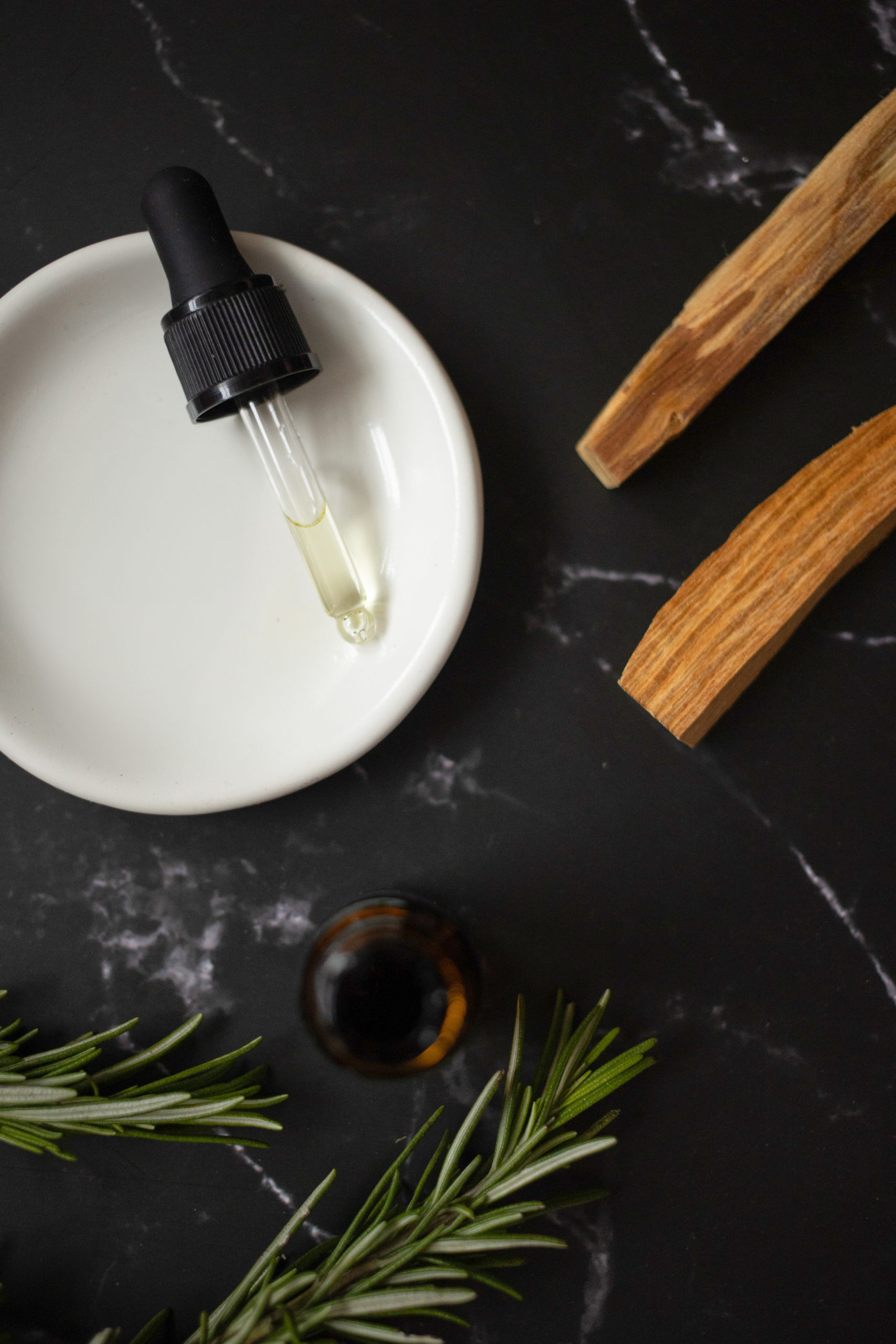 A dropper bottle with clear liquid rests on a small white dish, surrounded by green rosemary sprigs and wooden pieces, all on a dark marbled surface.