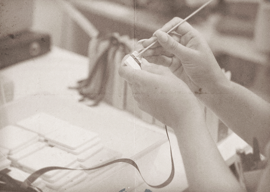 A person holds a small object while carefully applying paint with a brush, surrounded by various crafting materials. The image has a vintage, sepia tone.