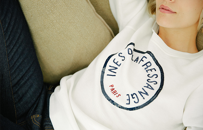 A close-up of a person wearing a white sweatshirt with a circular graphic that reads "Ines de la Fressange Paris," seated casually on a beige surface.