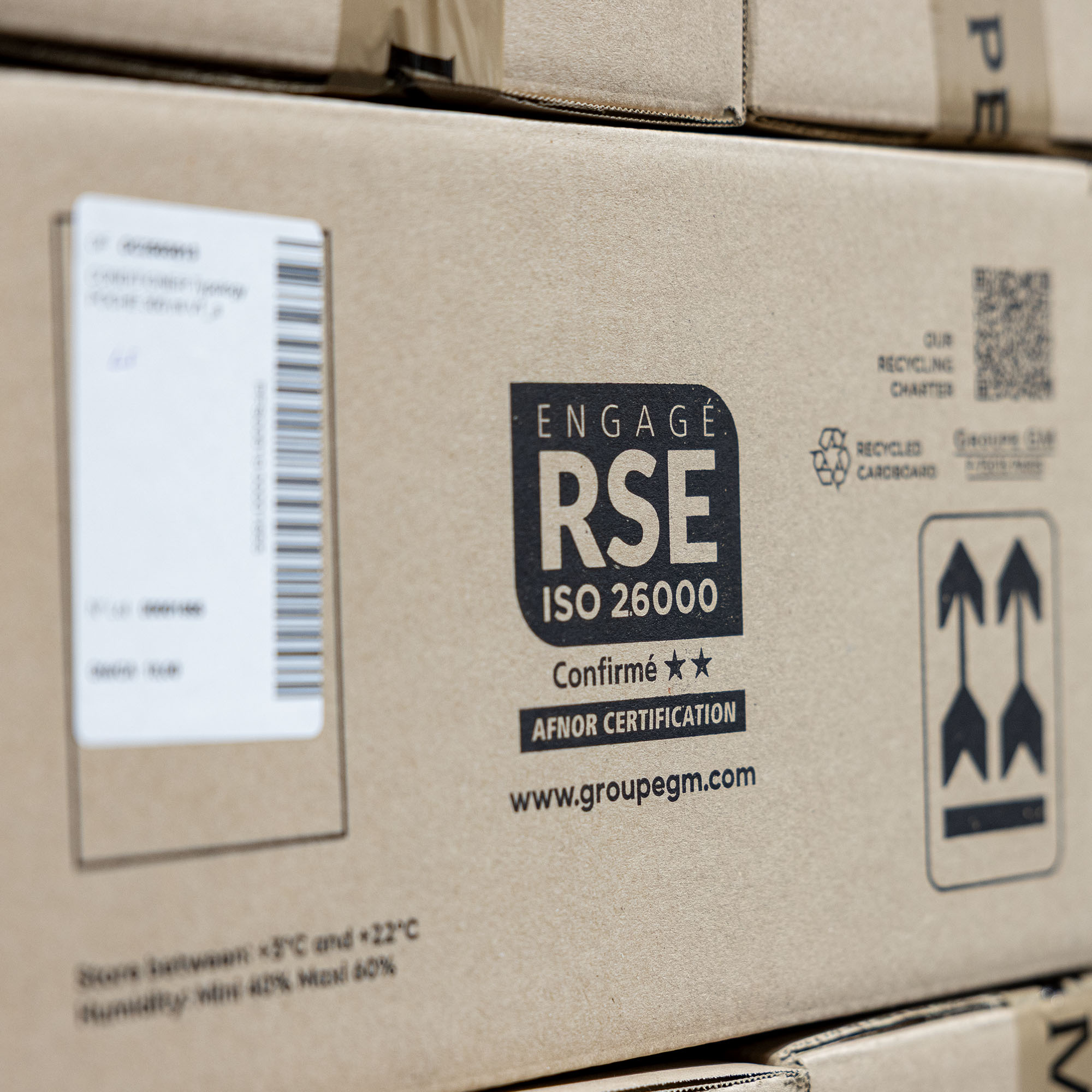 A close-up of a cardboard box displaying certifications and recycling information, with a focus on "ENGAGE RSE ISO 26000" and other related logos.