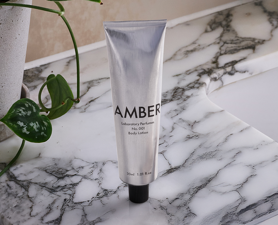 Un tubetto argentato di lozione per il corpo con l'etichetta "AMBER Laboratory Perfume No. 001" posizionato su una superficie di marmo con una pianta in vaso sullo sfondo.