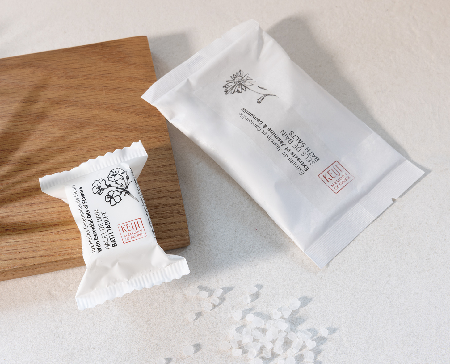 Two elegant packages of skincare products lie on a light background, accompanied by a scattering of coarse salt. One package is wrapped in white with botanical illustrations, while the other has a minimalist design featuring a daisy.