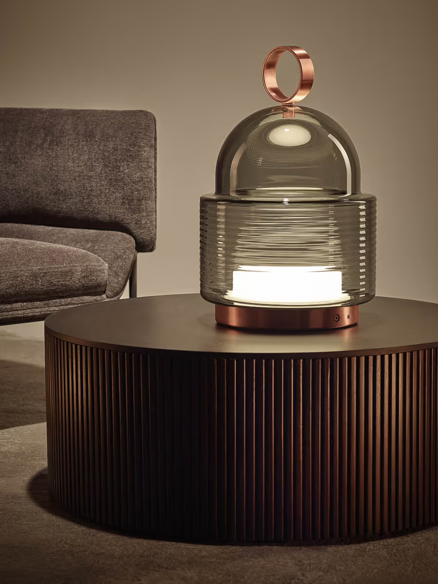 A modern Brokis lamp on a round, ribbed coffee table beside a gray couch.