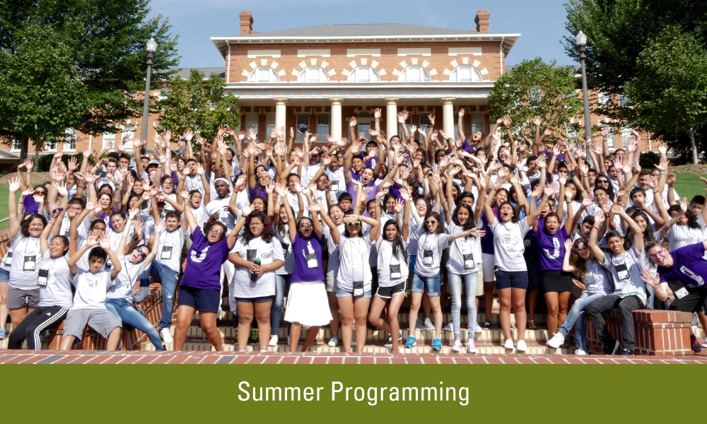 A group of adults and youth wearing white and purple shirts pose for a picture with their hands up to the sky on the steps of Court of Carolina. 