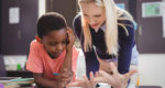 A teacher leans onto a desk next to a student and helps him understand a concept on his assignment.