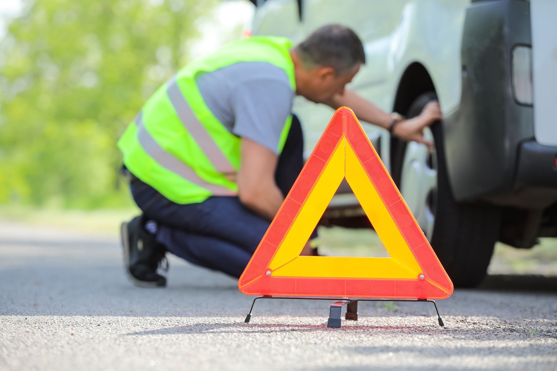 Man in safety vest checks tire, warning triangle on road.