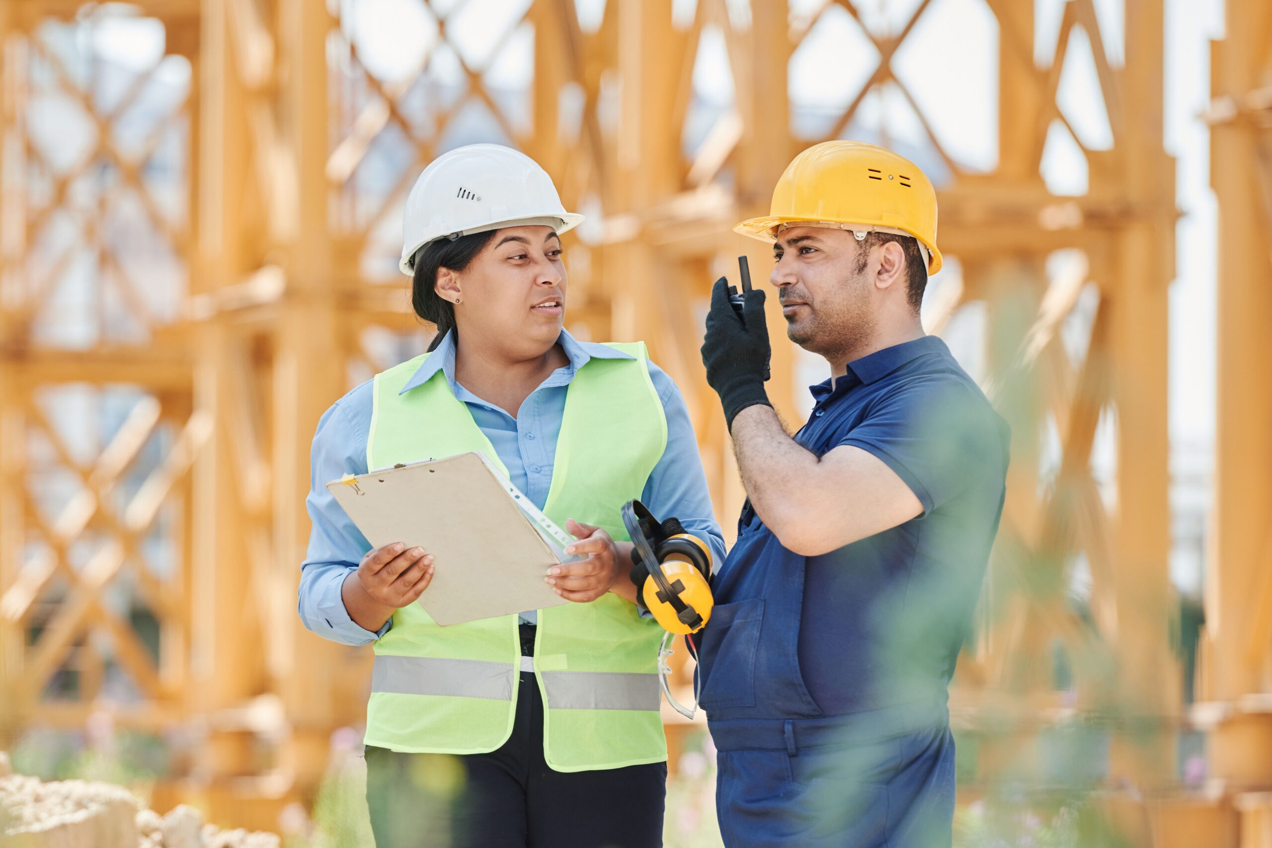 pexels-mikael-blomkvist-8961028 - Master Builders Association Two construction workers discuss on site: a woman in a white hard hat and high-visibility vest with a clipboard, and a man in a yellow hard hat in the background.