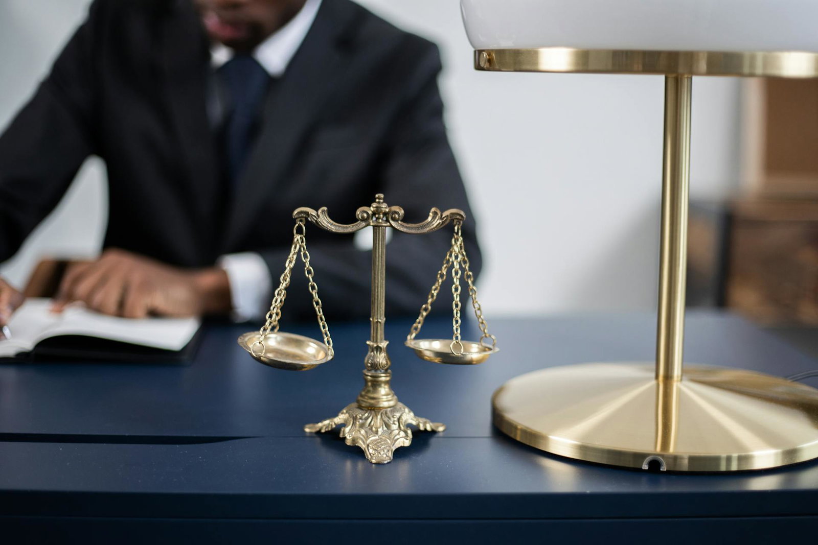 Brass scales of justice on a desk, symbolizing legal practice and professionalism, with a blurred figure of an attorney writing notes in the background.