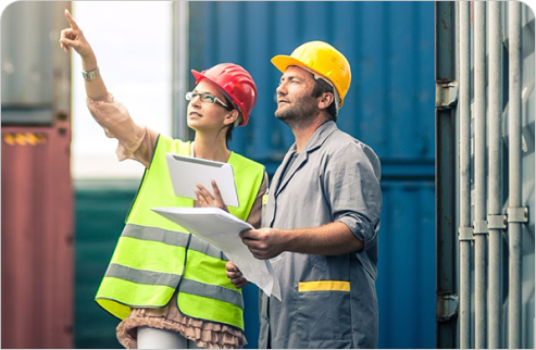 Two construction workers in safety helmets and vests reviewing plans at a shipping container site.