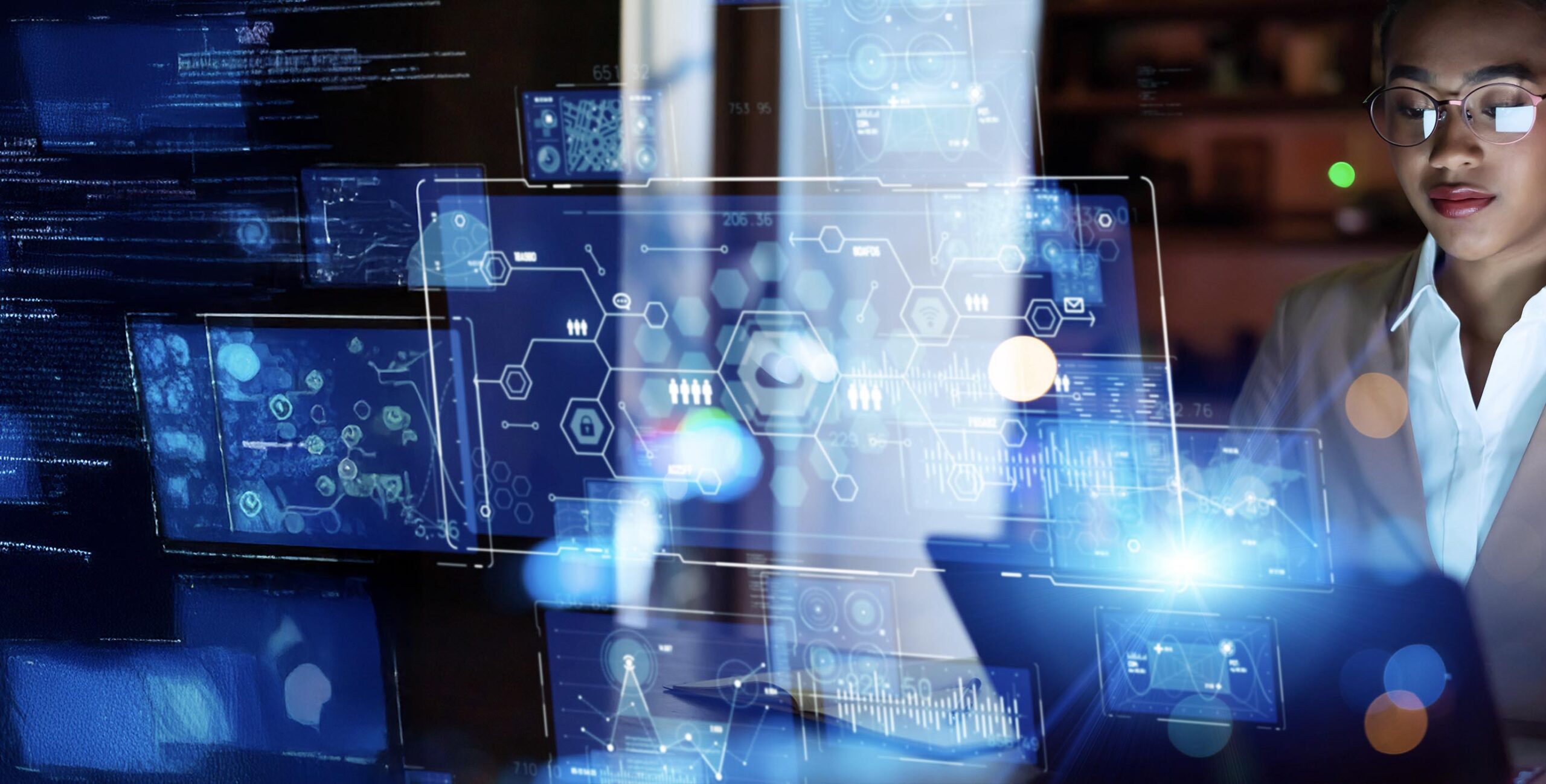 a professional looking woman in glasses looks down at a laptop, to the front of her glassy, digital images of data dashboards float in the air