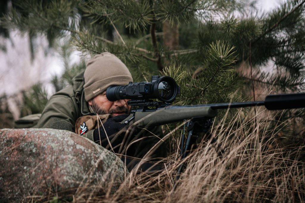 Jäger zielt mit einem Zielfernrohrgewehr aus der Deckung in grasbewachsenem Gelände