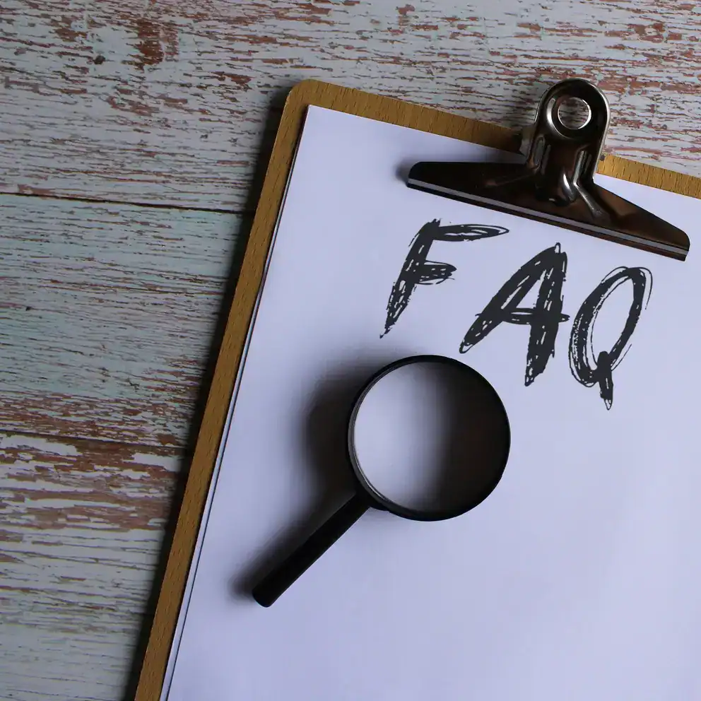 A clipboard holds a sheet of paper with "FAQ" written on it. A black magnifying glass rests on the paper, and the background is a wooden surface.