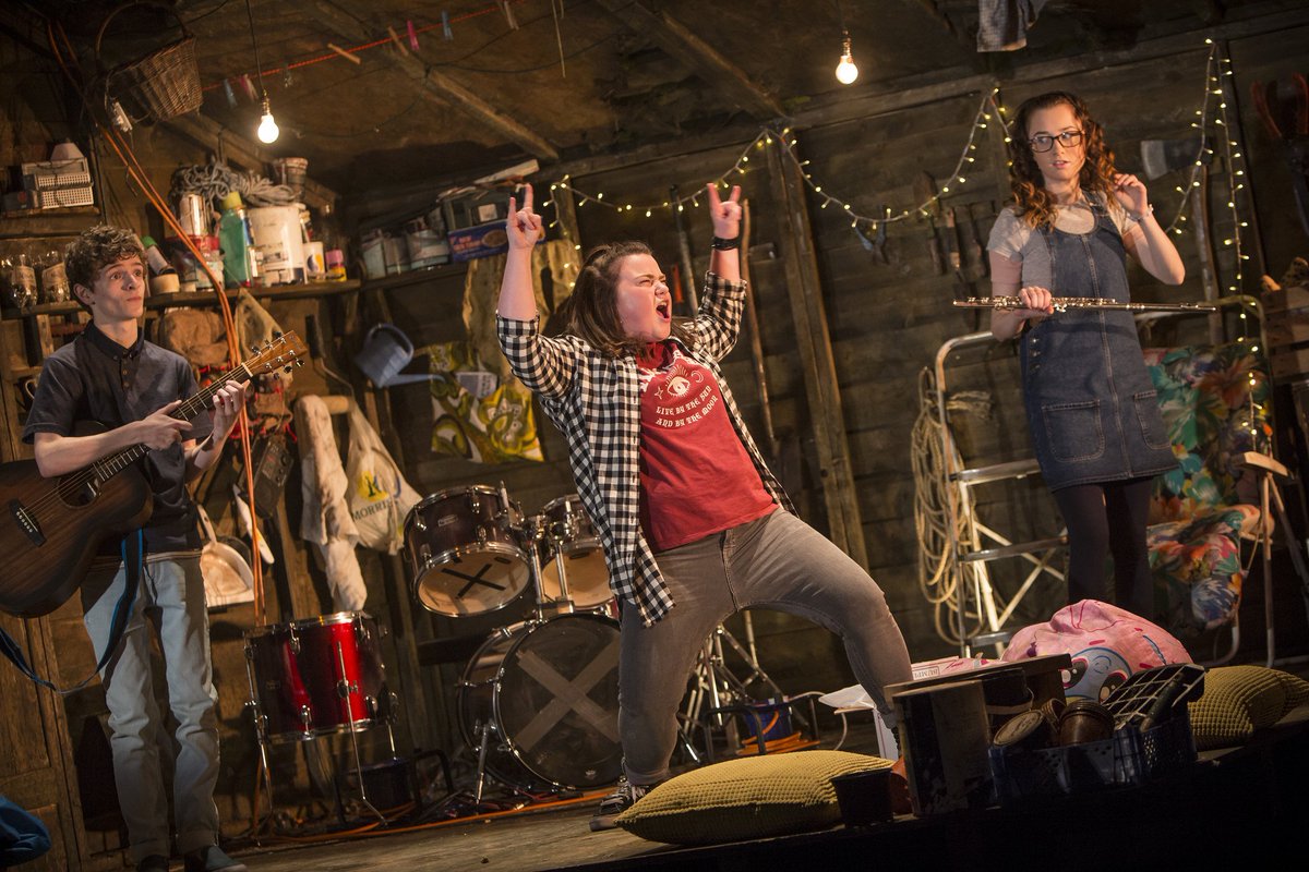 Three young people stand in a shed with musical instruments.