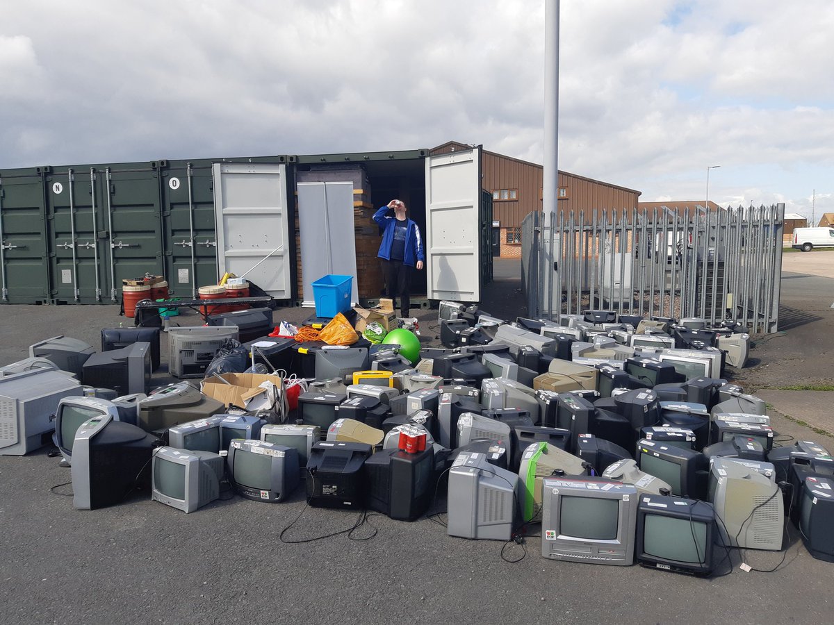 A storage container with well over 100 cathode ray televisions outside, and a northern man behind them, drinking a tin of beer.