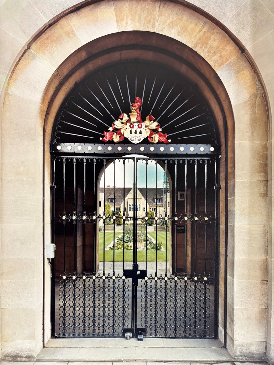 The Worcester Street gates, photo taken before College works started. A large iron gate into a curved archway, with a distant view of a long pond, green grass and College buildings through the entranceway.