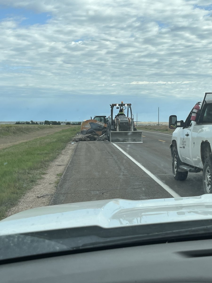 Image of construction vehicles on a road with a white truck parked nearby. The landscape is flat with grassy fields on either side and a cloudy sky overhead.