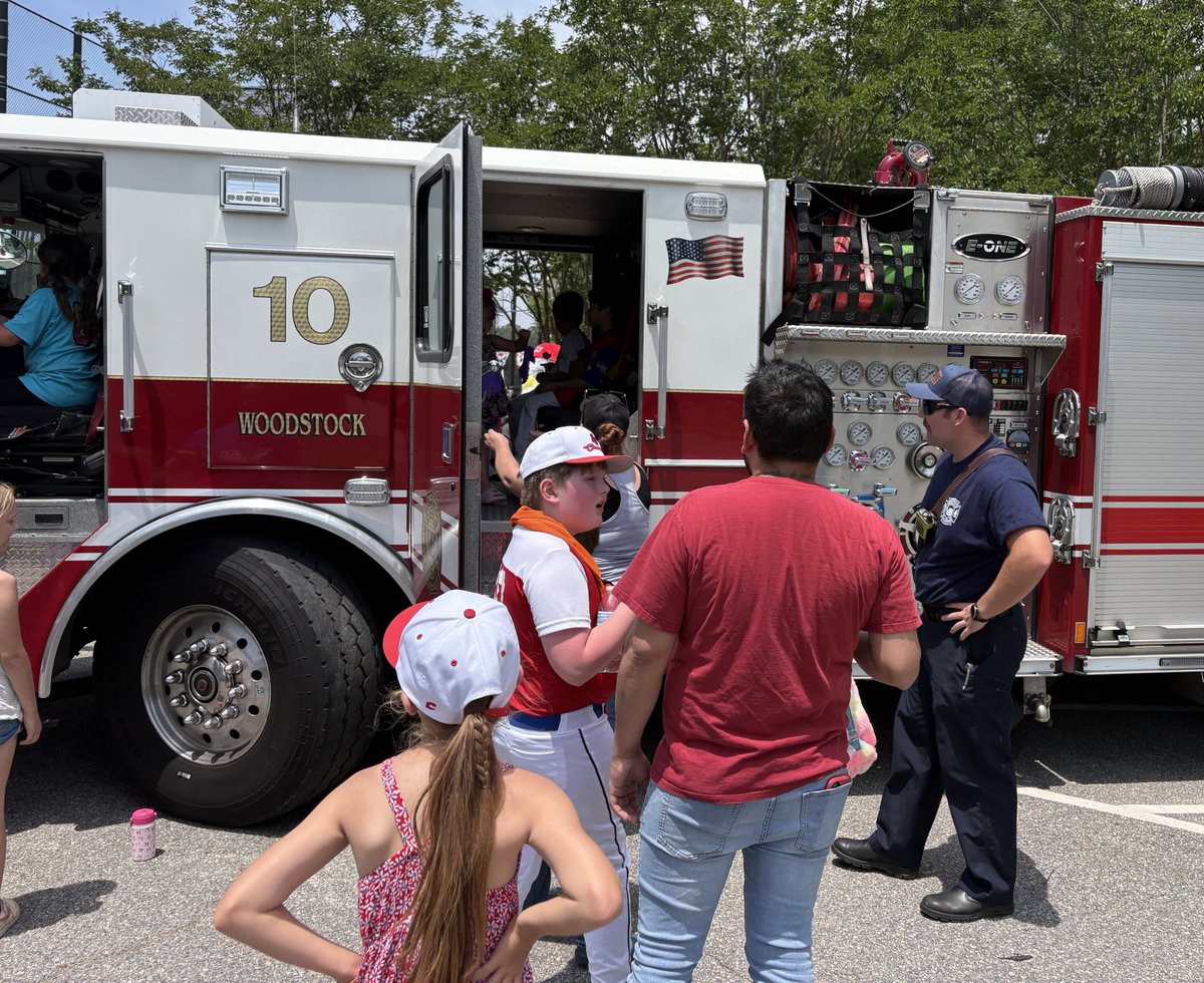 A group of people stands near a VBFD fire truck labeled "10." One person wearing a cap and uniform speaks with others. The truck has open compartments displaying equipment. Trees are visible in the background.