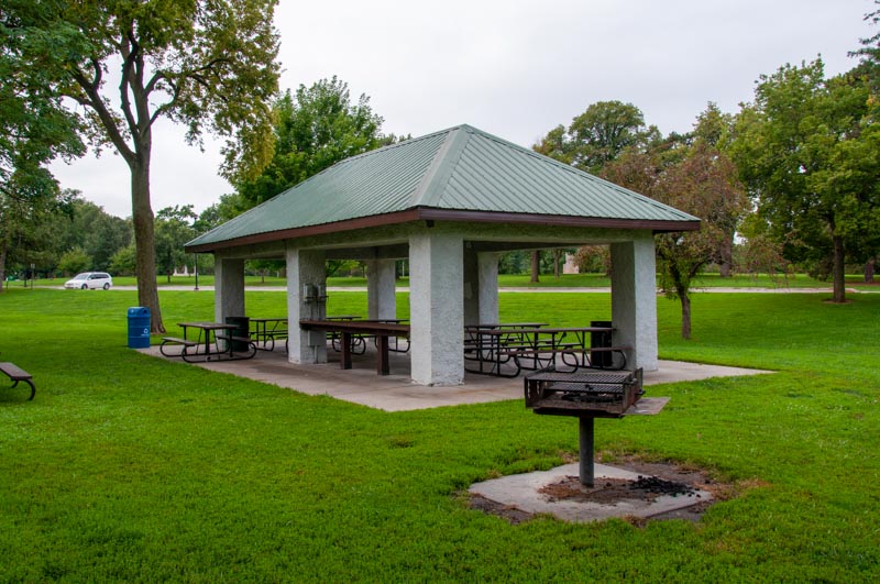 A park scene featuring a picnic pavilion with a green roof. Inside, there are several picnic tables. Nearby, there is an empty grill and a bench. 
