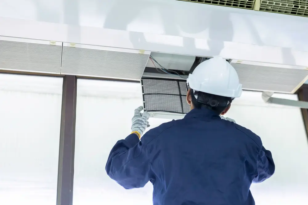 HVAC Technician Inspecting Ceiling-Mounted Air Conditioning Unit Filter HVAC technician in blue uniform and white hard hat removing or inspecting the air filter on a ceiling-mounted commercial air conditioning unit