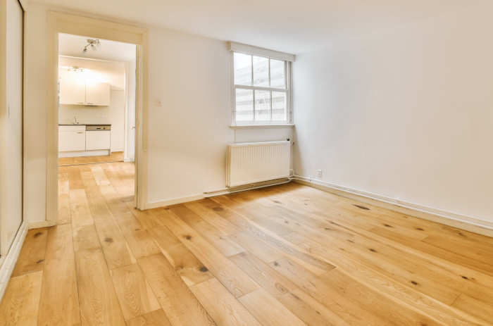 Empty room with wooden flooring leading to an adjoining kitchen area.