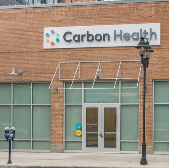 Storefront of a Carbon Health clinic with large windows, a brick exterior, and a company sign above the entrance. Two parking meters and a streetlamp are visible on the sidewalk.