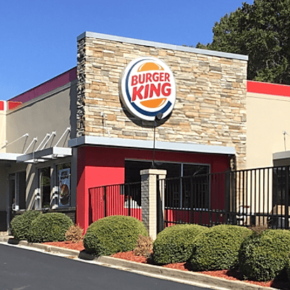 A Burger King restaurant with a stone facade, large logo sign, and landscaped bushes in front, owned by Carrols.