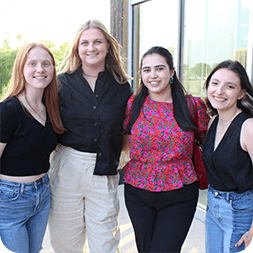 Four young women standing together outside, smiling at the camera, celebrate their internship in construction program management.