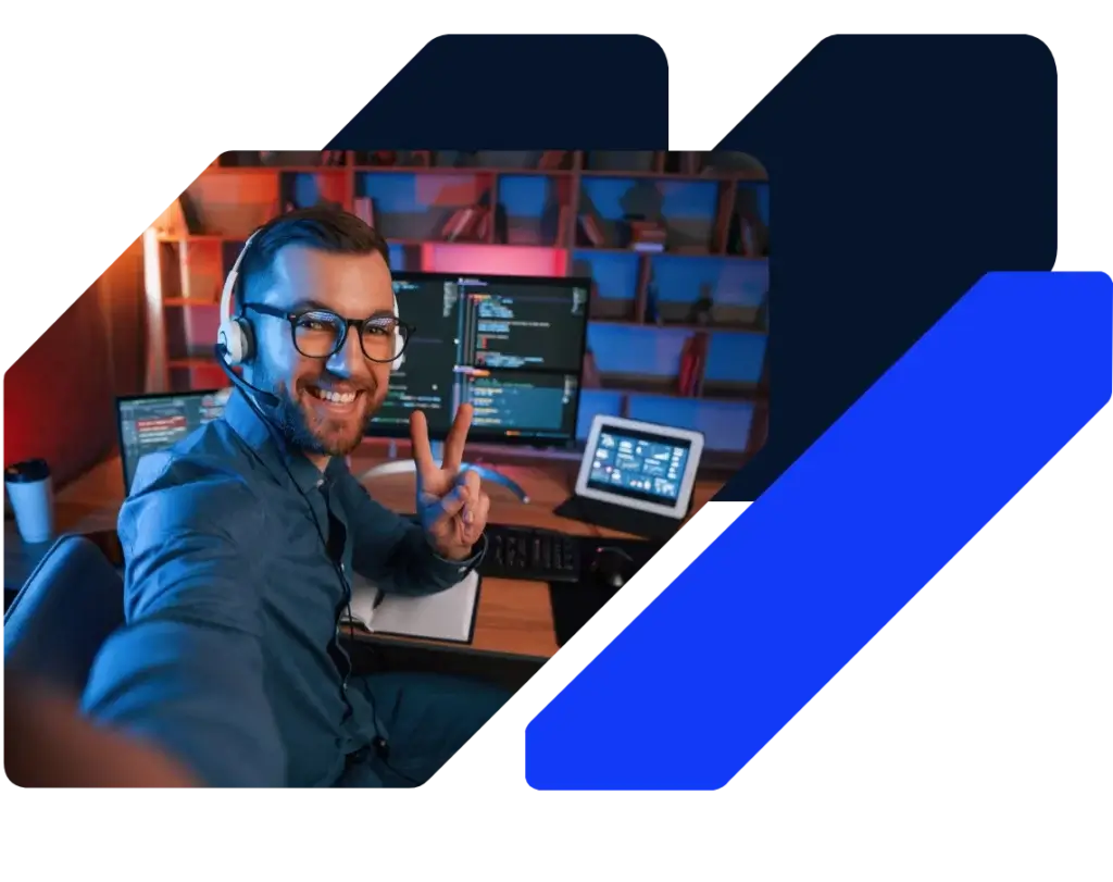 A smiling man with a headset sits at a computer desk, making a peace sign at the camera. Multiple monitors display web design code, and bookshelves are visible in the background. Blue graphic shapes overlay the image.