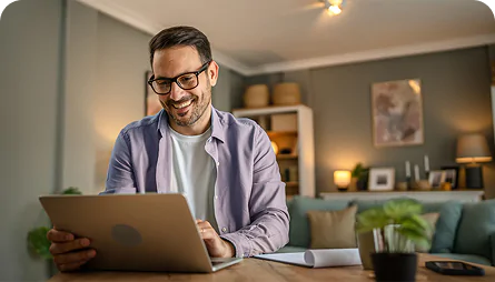 Ein lächelnder Mann mit Brille arbeitet an einem Laptop in einem gemütlichen Wohnzimmer. Auf dem Tisch liegen Unterlagen und eine kleine Topfpflanze; im Hintergrund ist ein Sofa zu sehen.